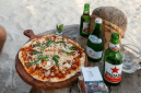 Dua botol bir Bintang, botol air mineral Aqua Panna, serta satu loyang pizza ukuran medium beralas papan kayu menghiasi meja kayu di pinggir Pantai Kuta, Lombok Tengah. (Foto: Lombokini.com/Tangkapan Layar)