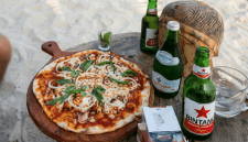 Dua botol bir Bintang, botol air mineral Aqua Panna, serta satu loyang pizza ukuran medium beralas papan kayu menghiasi meja kayu di pinggir Pantai Kuta, Lombok Tengah. (Foto: Lombokini.com/Tangkapan Layar)